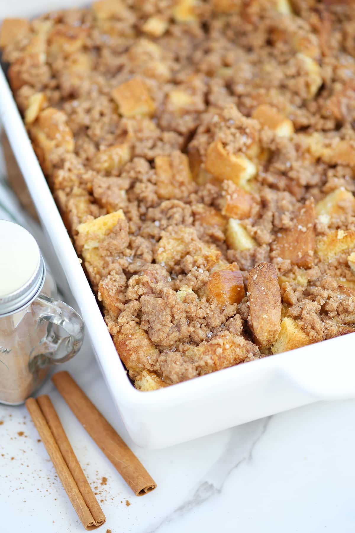 Baked French Toast in a white ceramic baking dish with cinnamon sticks and a cinnamon shaker in the background.