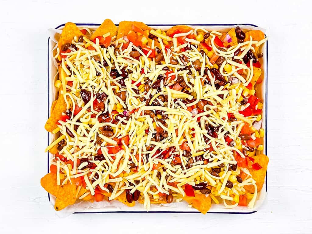 Chips topped with veggies, black beans and cheese on a white baking dish.